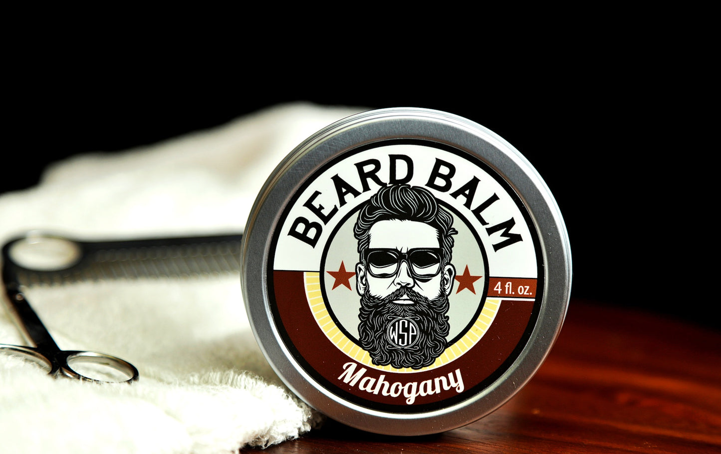Natural beard balm in stylish tin container on a wooden table with a towel, scissors, and a metal comb. Handcrafted beard balm made in small batches in America. Beard balm ingredients including Shea Butter, Jojoba Oil, and Avocado Oil. Beard leave-in conditioner. Mahogany scented beard balm.