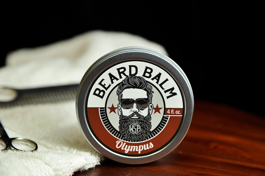 Natural beard balm in stylish tin container on a wooden table with a towel, scissors, and a metal comb. Handcrafted beard balm made in small batches in America. Beard balm ingredients including Shea Butter, Jojoba Oil, and Avocado Oil. Beard leave-in conditioner. Olympus scented beard balm.