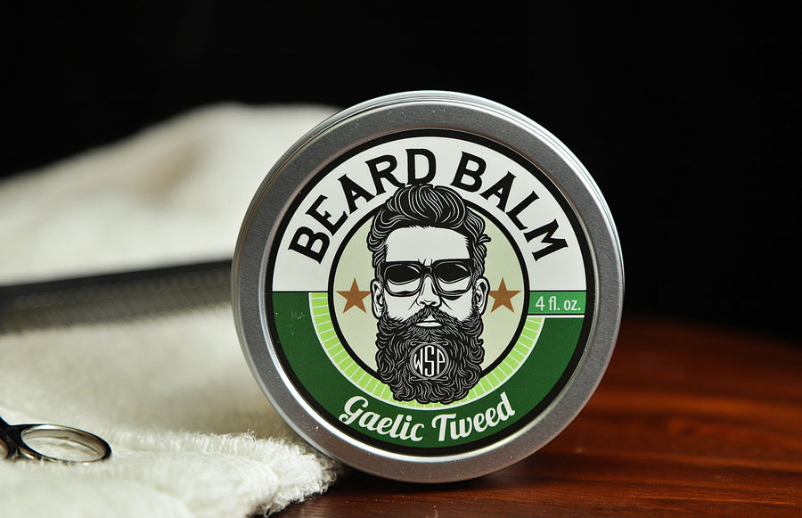 Natural beard balm in stylish tin container on a wooden table with a towel, scissors, and a metal comb. Handcrafted beard balm made in small batches in America. Beard balm ingredients including Shea Butter, Jojoba Oil, and Avocado Oil. Beard leave-in conditioner. Gaelic tweed scented beard balm.