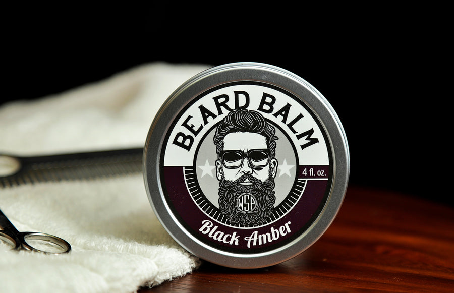 Natural beard balm in stylish tin container on a wooden table with a towel, scissors, and a metal comb. Handcrafted beard balm made in small batches in America. Beard balm ingredients including Shea Butter, Jojoba Oil, and Avocado Oil. Beard leave-in conditioner.  Black Amber Vanille Scented beard balm. Black Amber.
