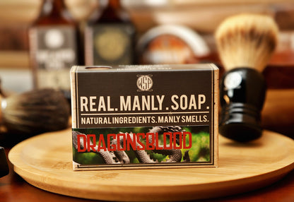 Bar of soap with 'Real Manly Soap' Dragonsblood scent packaging on a wooden tray, surrounded by shaving tools.