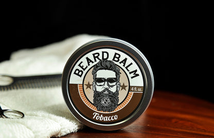 Natural beard balm in stylish tin container on a wooden table with a towel, scissors, and a metal comb. Handcrafted beard balm made in small batches in America. Beard balm ingredients including Shea Butter, Jojoba Oil, and Avocado Oil. Beard leave-in conditioner. Tobacco scented beard balm.