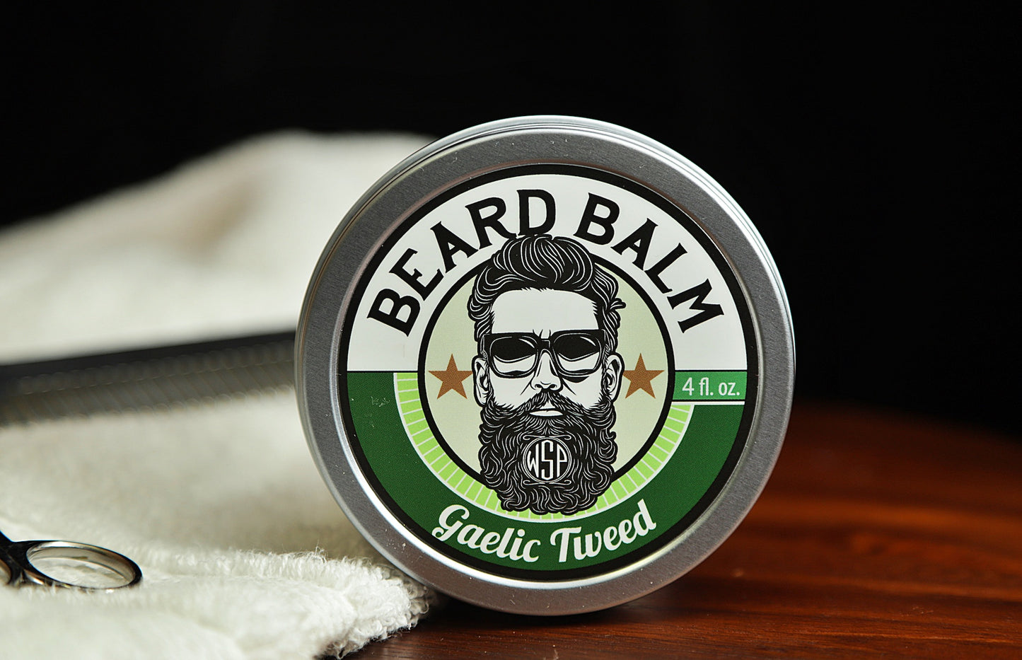 Natural beard balm in stylish tin container on a wooden table with a towel, scissors, and a metal comb. Handcrafted beard balm made in small batches in America. Beard balm ingredients including Shea Butter, Jojoba Oil, and Avocado Oil. Beard leave-in conditioner. Gaelic tweed scented beard balm.