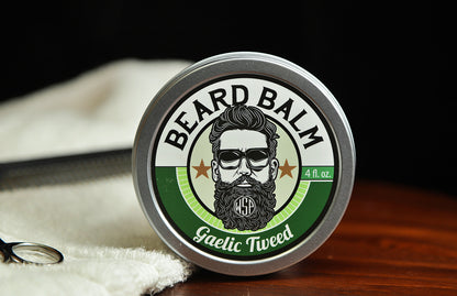 Natural beard balm in stylish tin container on a wooden table with a towel, scissors, and a metal comb. Handcrafted beard balm made in small batches in America. Beard balm ingredients including Shea Butter, Jojoba Oil, and Avocado Oil. Beard leave-in conditioner. Gaelic tweed scented beard balm.