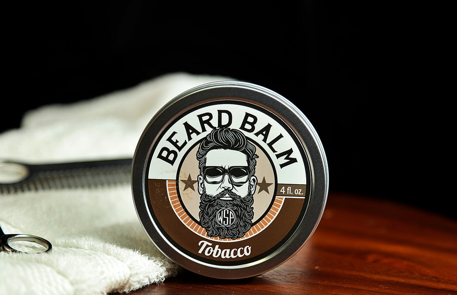 Natural beard balm in stylish tin container on a wooden table with a towel, scissors, and a metal comb. Handcrafted beard balm made in small batches in America. Beard balm ingredients including Shea Butter, Jojoba Oil, and Avocado Oil. Beard leave-in conditioner. Tobacco scented beard balm.