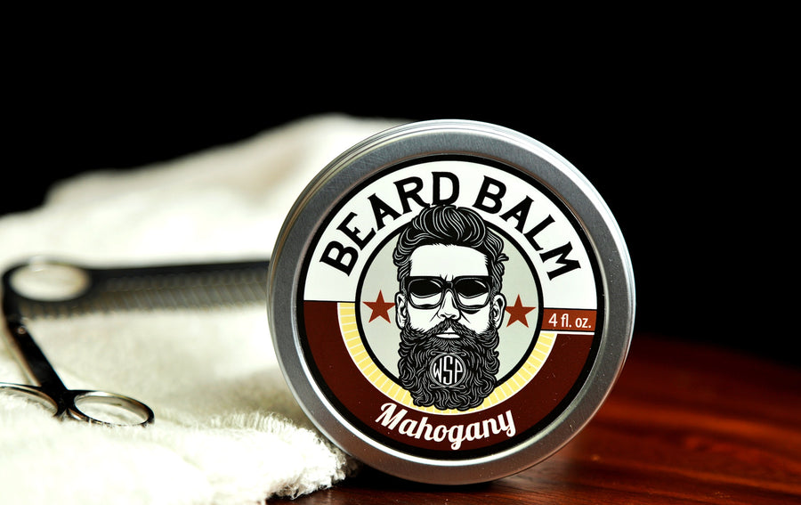 Natural beard balm in stylish tin container on a wooden table with a towel, scissors, and a metal comb. Handcrafted beard balm made in small batches in America. Beard balm ingredients including Shea Butter, Jojoba Oil, and Avocado Oil. Beard leave-in conditioner.  Mahogany scented beard balm.