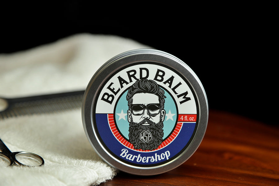 Natural beard balm in stylish tin container on a wooden table with a towel, scissors, and a metal comb. Handcrafted beard balm made in small batches in America. Beard balm ingredients including Shea Butter, Jojoba Oil, and Avocado Oil. Beard leave-in conditioner. Barbershop Scented beard balm.