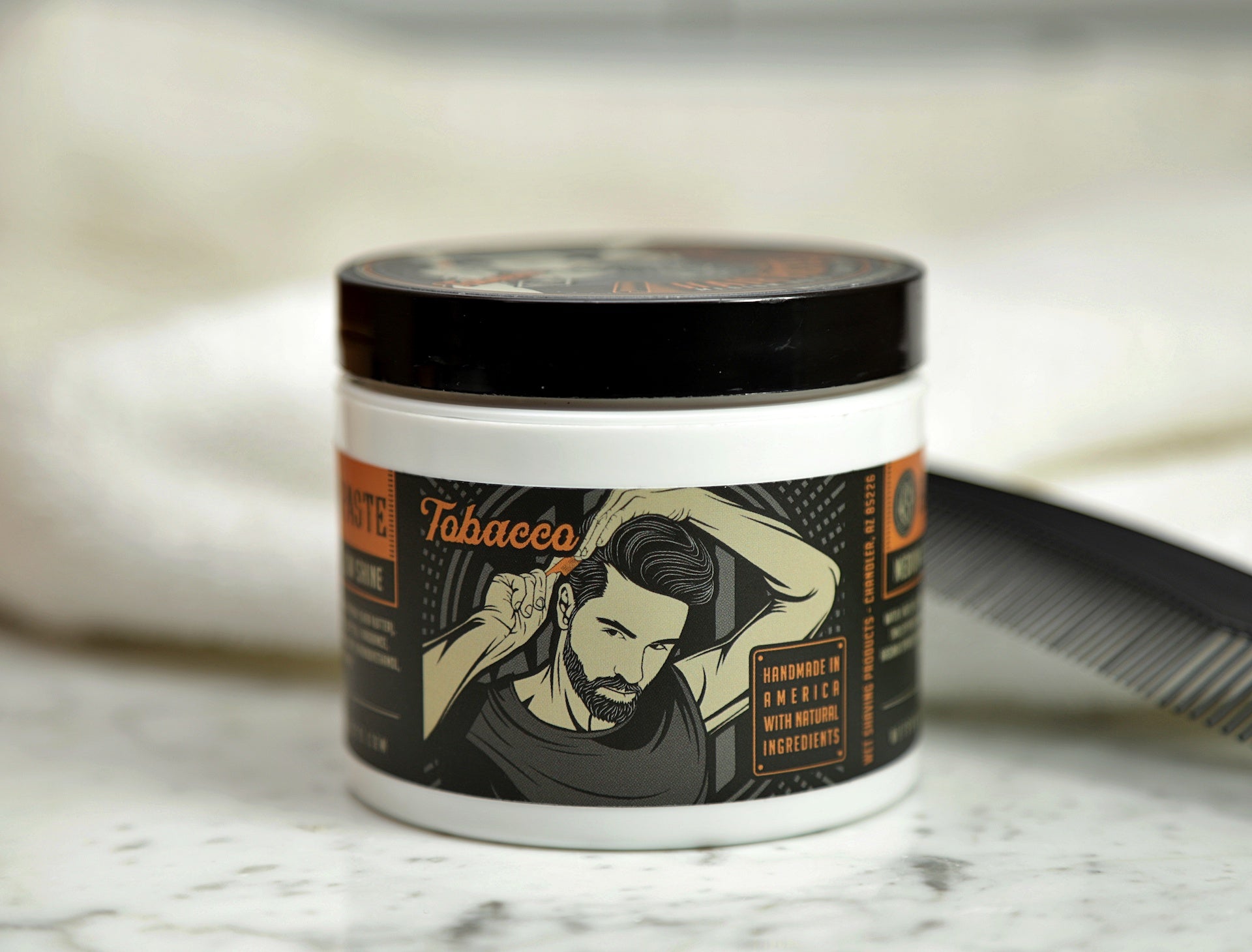 Front view of hair paste tub in Tobacco Scent with a comb and towel set on a marble counter